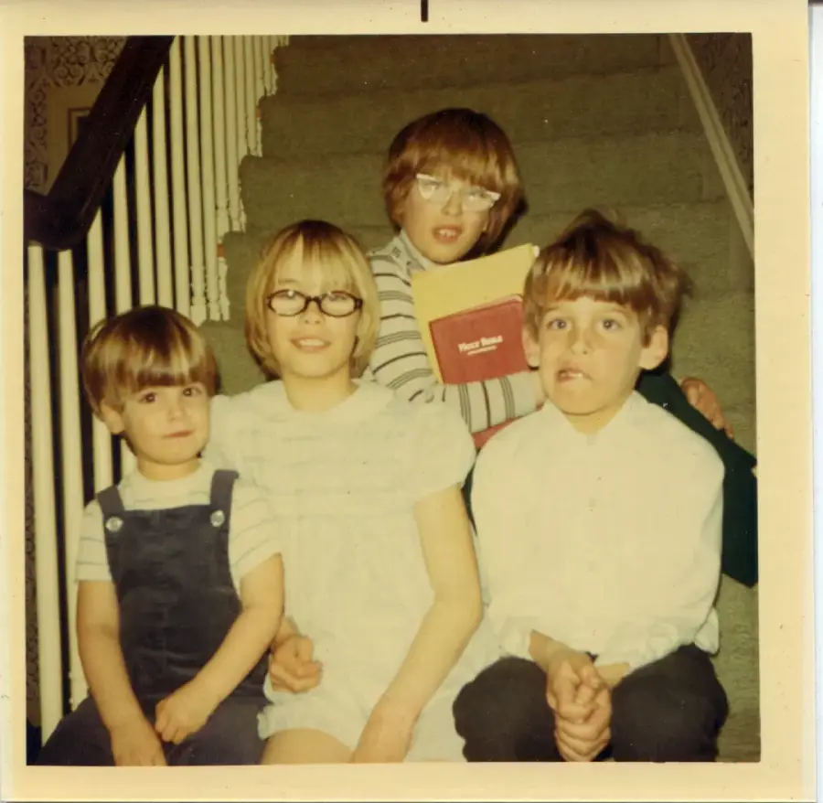 Four children sit together on a carpeted staircase, smiling at the camera. One child holds a red book. The children wear casual clothing and glasses, and a wooden railing is visible on the left side.