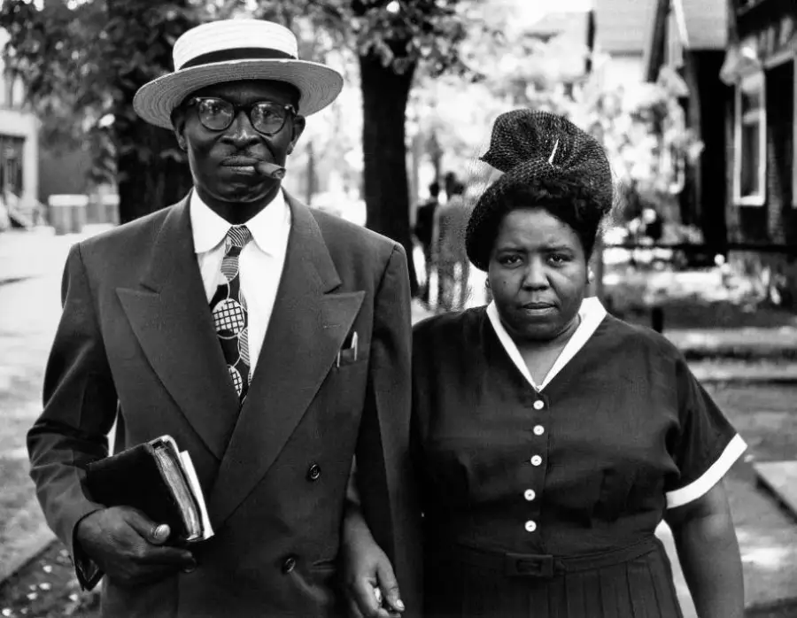 A man in a suit, hat, and glasses holding a book and a woman in a dress with a head wrap stand side by side outdoors on a sidewalk, both looking at the camera.