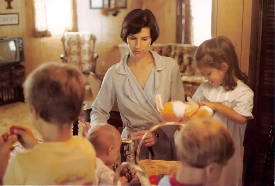 A woman stands in a cozy, softly lit living room, surrounded by three children and a baby, as they look through baskets and toys together. The scene feels warm and family-oriented.