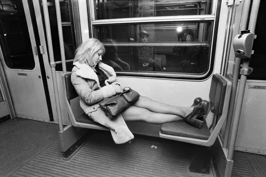 A woman in a coat and heels sleeps on a subway bench, her legs stretched out across the seats, with her purse in her lap. The subway car appears empty, with her reflection visible in the window.