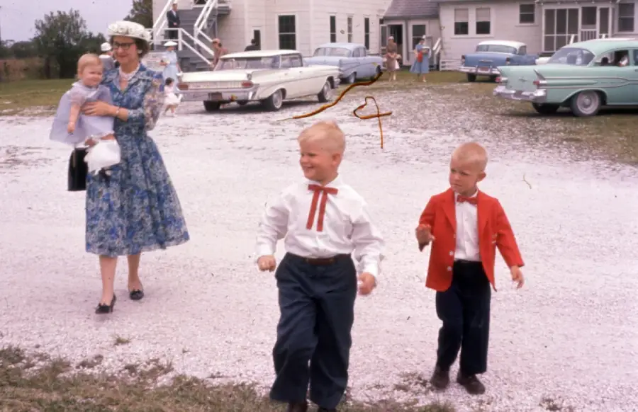 A woman in a blue floral dress and hat holds a baby while walking on gravel with two young boys; one boy wears a white shirt with a red tie, the other a red jacket. Vintage cars and a white building are in the background.