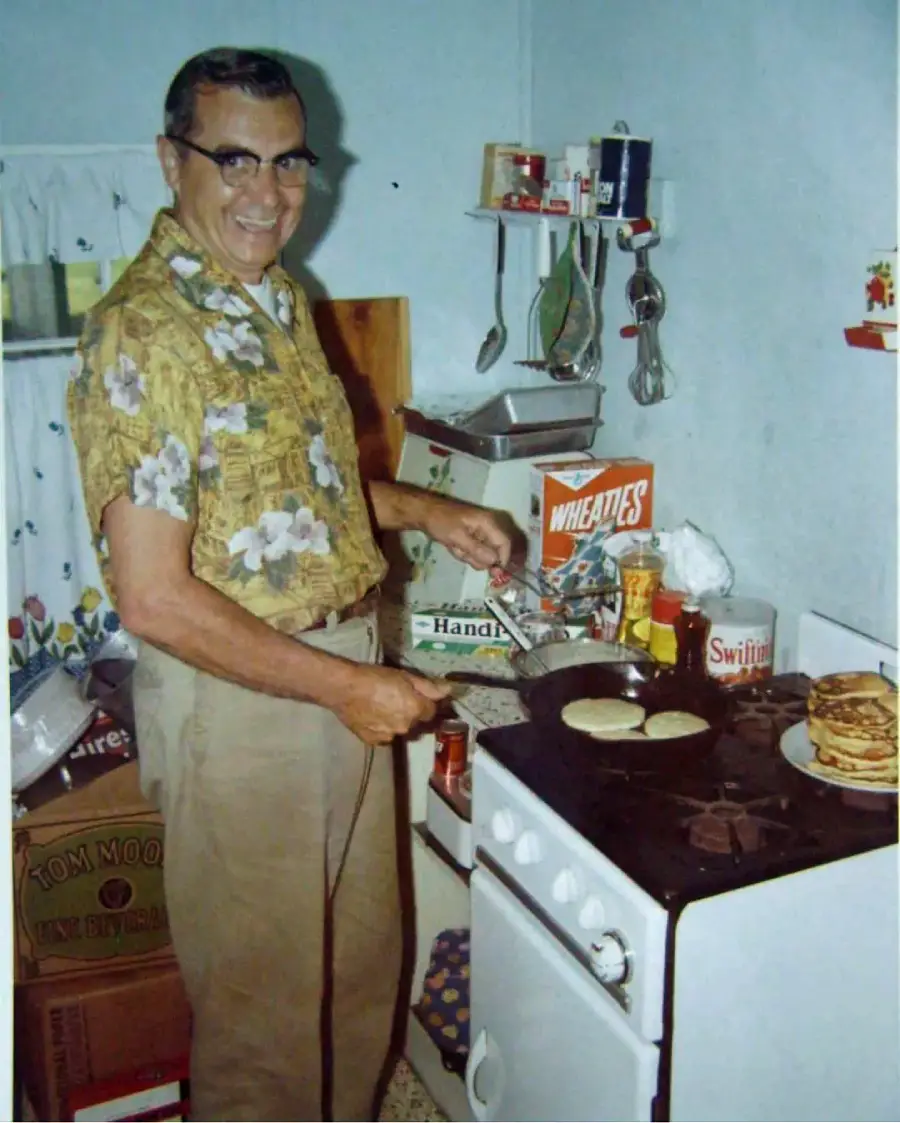 A man in glasses and a floral shirt smiles while flipping pancakes on a stove in a modest kitchen. Pancake mix, syrup, and a box of Wheaties are on the counter; stacked pancakes are visible beside him.