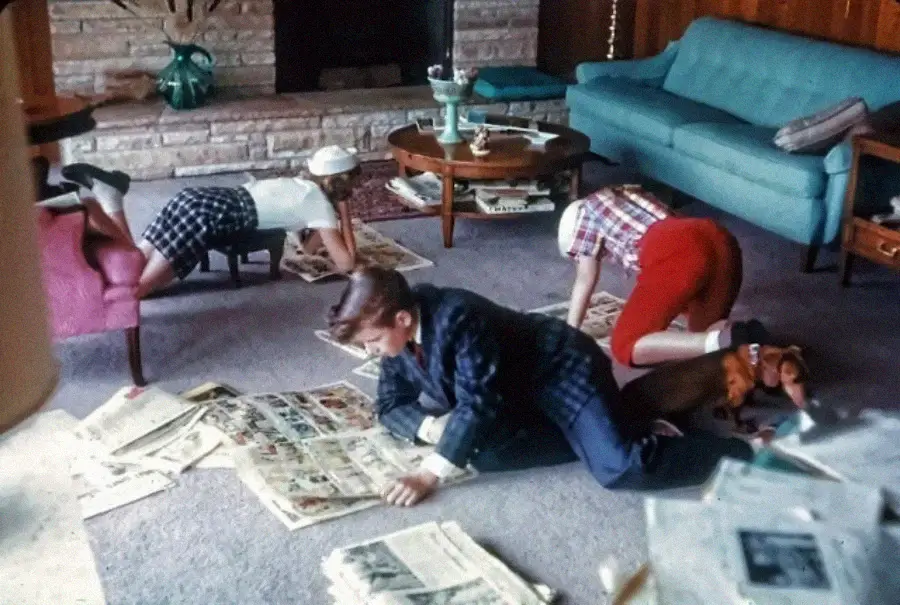 Three children and a dog are on the living room floor, intently reading comic sections of newspapers spread out on the carpet. The room has a blue couch, coffee table, and stone fireplace in the background.