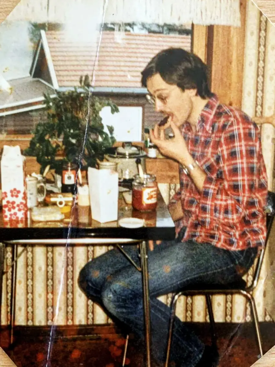 A person in a red plaid shirt and jeans sits at a table by a window, eating while surrounded by food items, mugs, and a plant in a cozy, vintage-style room.
