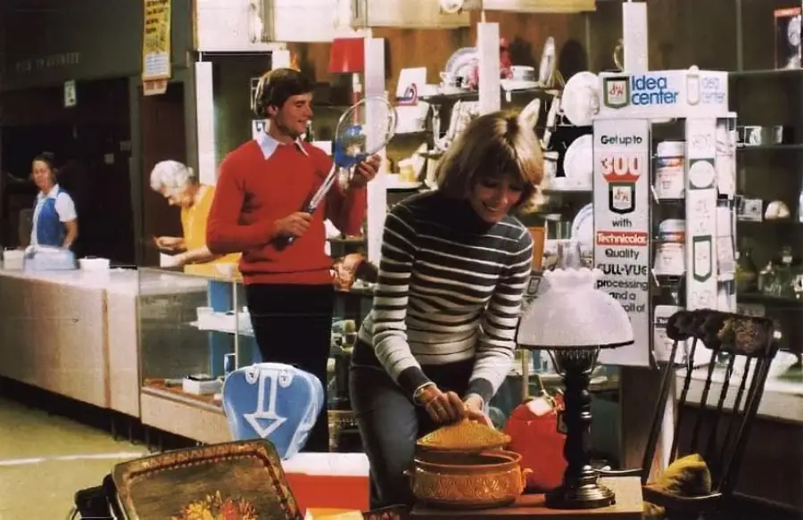 A woman in a striped sweater examines a basket near a lamp and chair in a store, while a man in a red sweater holds a racket behind her; shelves of electronics and appliances are visible in the background.
