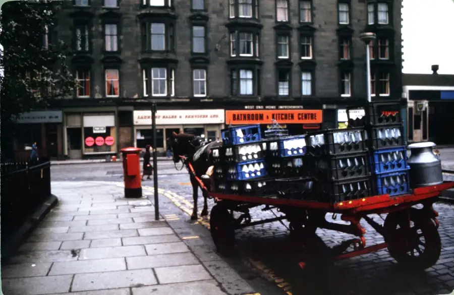 A horse-drawn cart loaded with crates and a milk can is parked beside a sidewalk on a city street, with shops and apartments visible in the background.