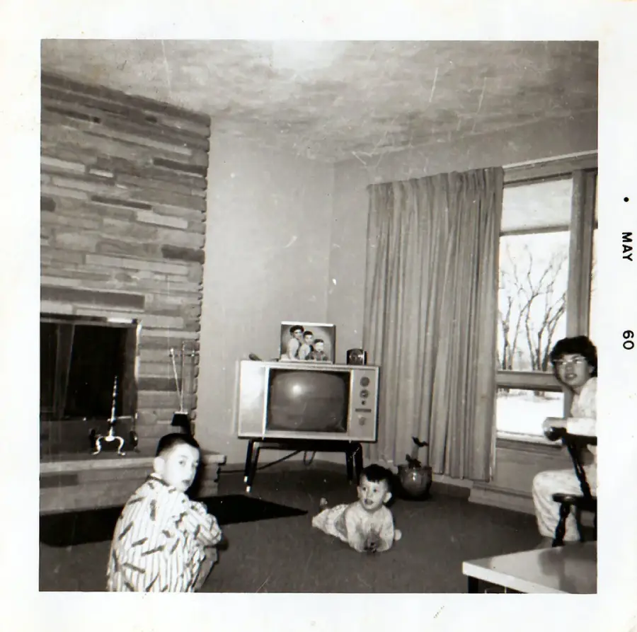 Black and white photo from May 1960 showing two children and an adult in pajamas in a living room with a fireplace, vintage TV, potted plant, large curtained window, and family photos on the TV.