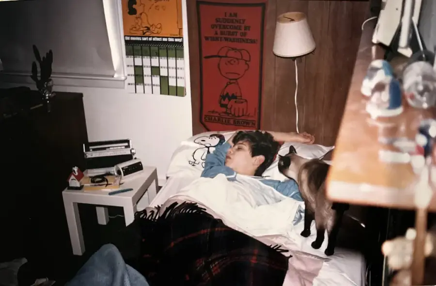 A boy sleeps in a bed with Snoopy sheets while a Siamese cat stands nearby. A Charlie Brown poster and a calendar hang on the wall above the bed. The room has a lamp, books, and small figurines.
