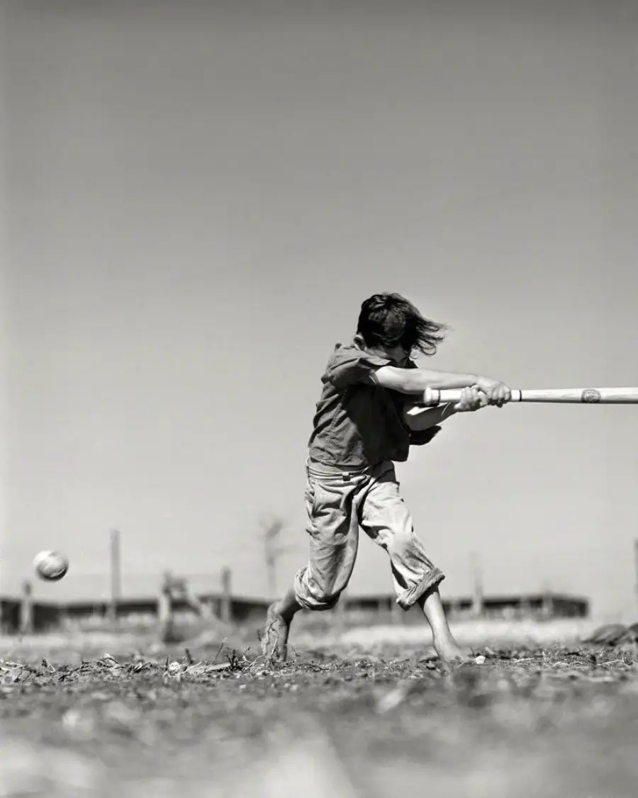 A barefoot child with windswept hair swings a baseball bat at an incoming ball outdoors on a dirt field, with blurred buildings and a clear sky in the background.