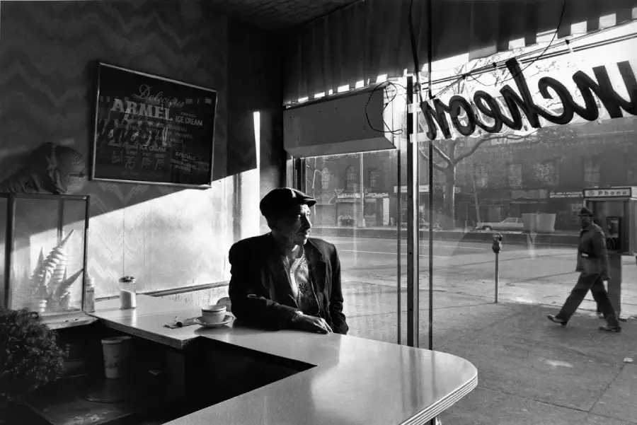 An older man in a cap sits alone at a diner counter by a large window, with sunlight streaming in. A coffee cup is on the counter, and the street outside is visible through the glass, along with a passing pedestrian.