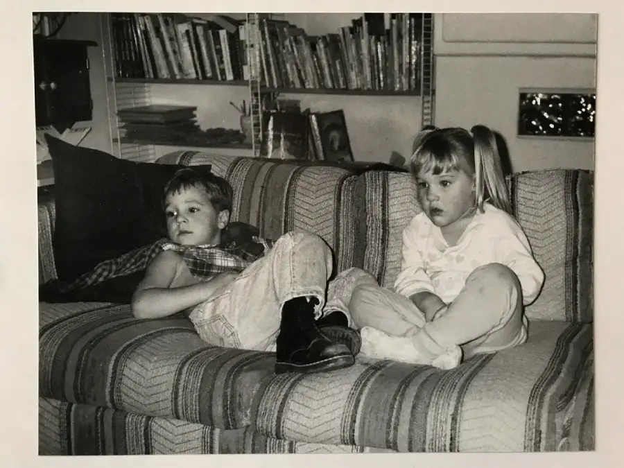 Two young children sit on a striped couch. The boy on the left is lounging with his shirt half off, and the girl on the right sits with her legs crossed. A bookshelf and window appear in the background. The image is black and white.