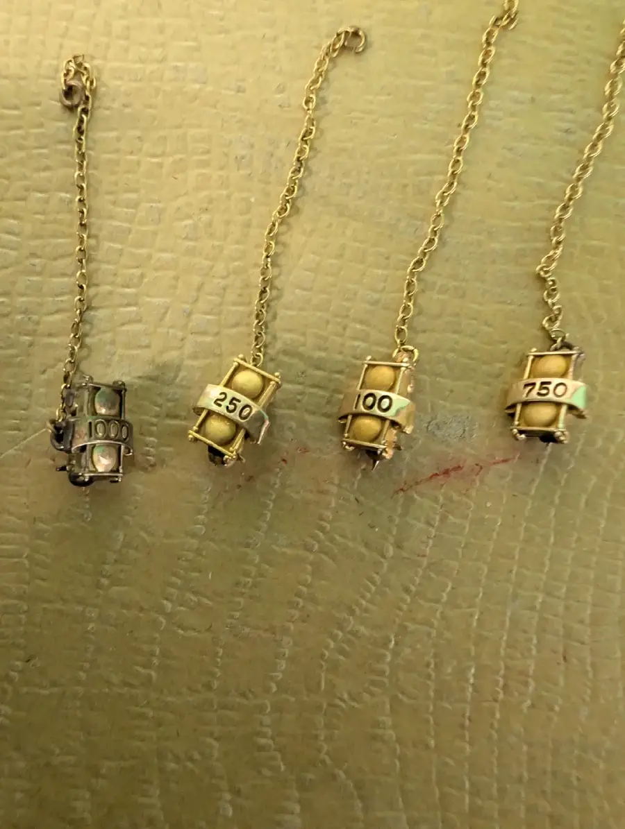 Four gold chain necklaces, each with a cylindrical pendant featuring a number—100, 250, 100, and 750—displayed on a light, textured surface.
