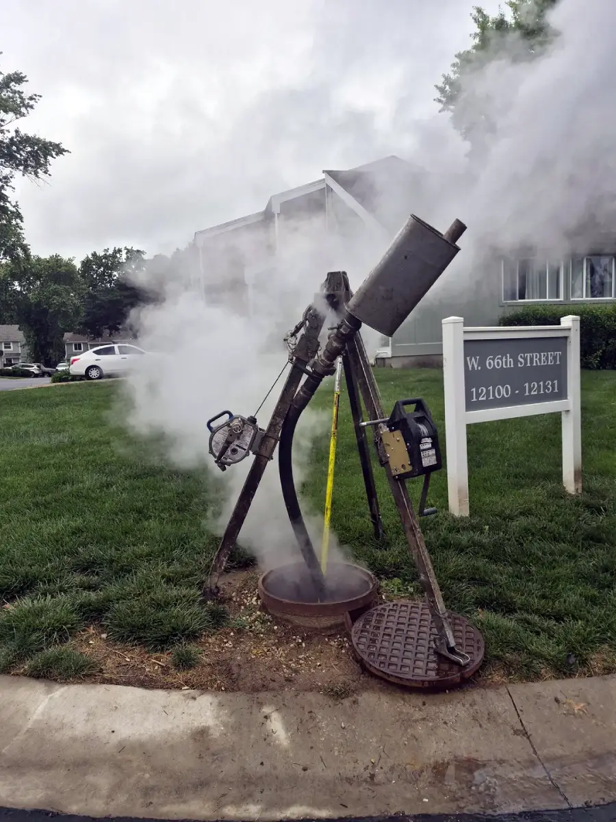 A large machine emitting white steam stands over an open manhole on a grassy area next to a sidewalk, with a street sign for W. 66th Street and nearby buildings visible in the background.