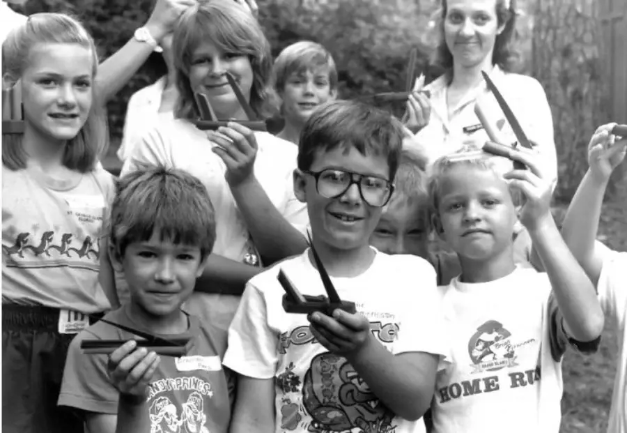 A group of smiling children and adults pose outdoors, holding small wooden model airplanes. The children stand in the front, some wearing graphic T-shirts, and everyone looks happy and excited.