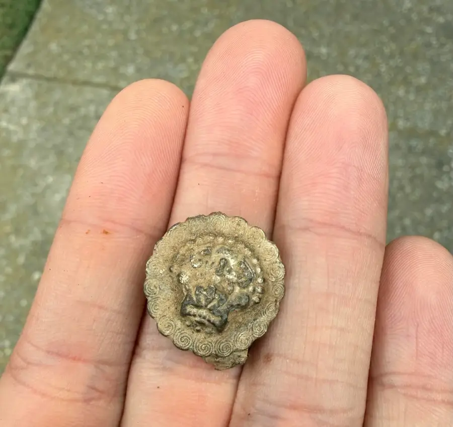 A close-up of a hand holding a small, round, weathered, ornamental object with scalloped edges and a faint, worn design in the center. The background shows a concrete surface and some grass.