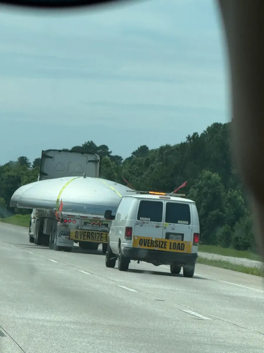 A large truck transports a wind turbine blade secured on its trailer, accompanied by a white van with an "Oversize Load" sign, driving on a multi-lane highway surrounded by trees.