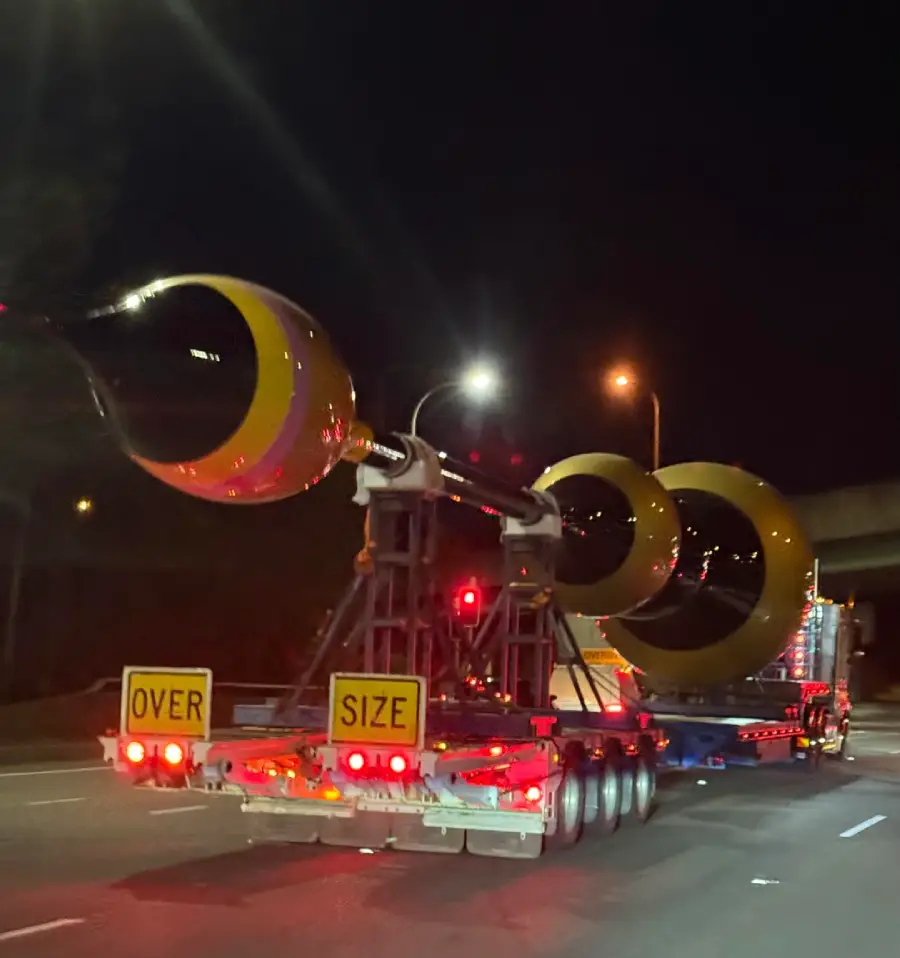 A large flatbed truck transports an oversized, shiny black and yellow object with a rounded, pointed end at night. The object is securely fastened, and "OVER SIZE" signs are displayed on the truck's rear.