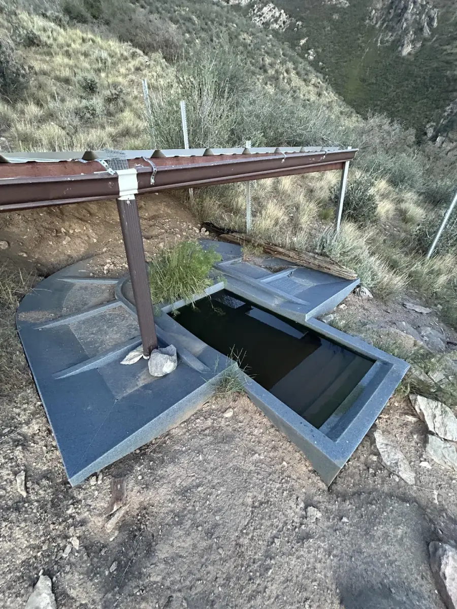 A small, rectangular concrete water trough is built into a hillside, collecting water from a brown gutter system above. Some grass grows beside the trough, and the area is surrounded by rocks and dry, grassy terrain.