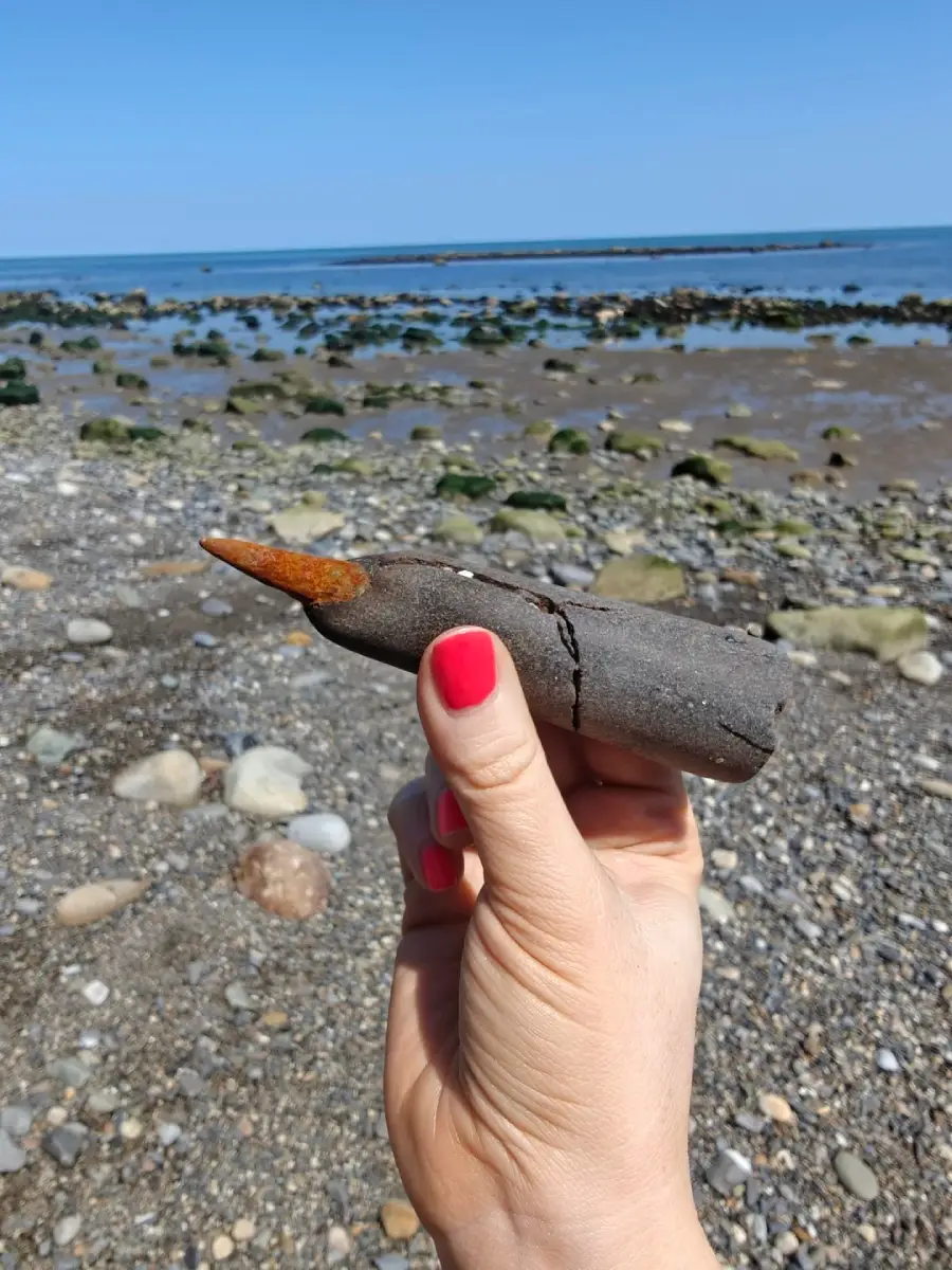 A hand with red-painted nails holds a large, rusty bullet-shaped object on a rocky beach with the sea and blue sky in the background.