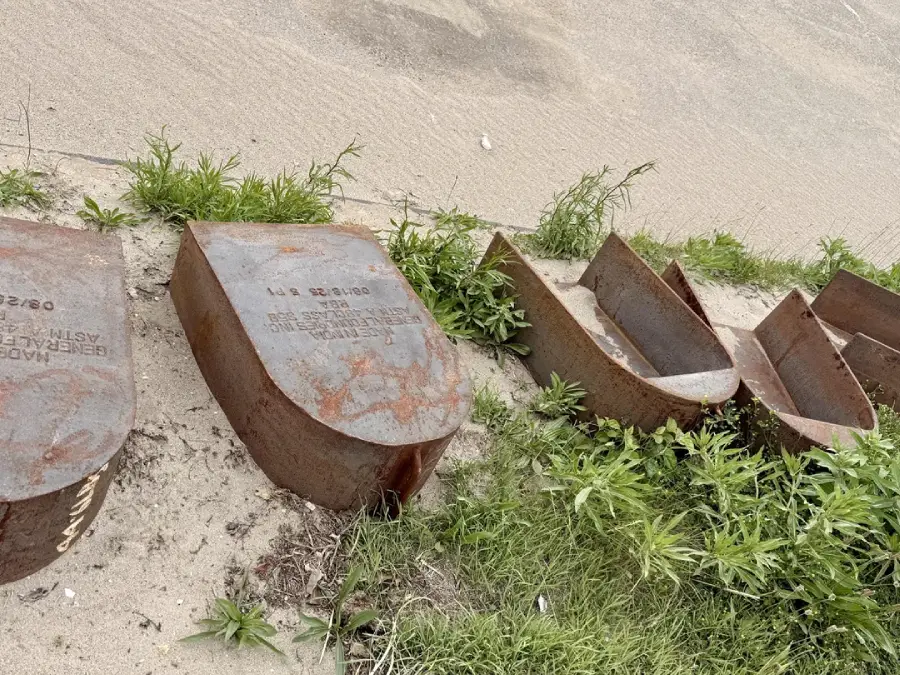 Six large, rusted metal wedges partially buried in sand and surrounded by patches of green grass near a concrete surface. Some stamped markings are visible on the metal pieces.