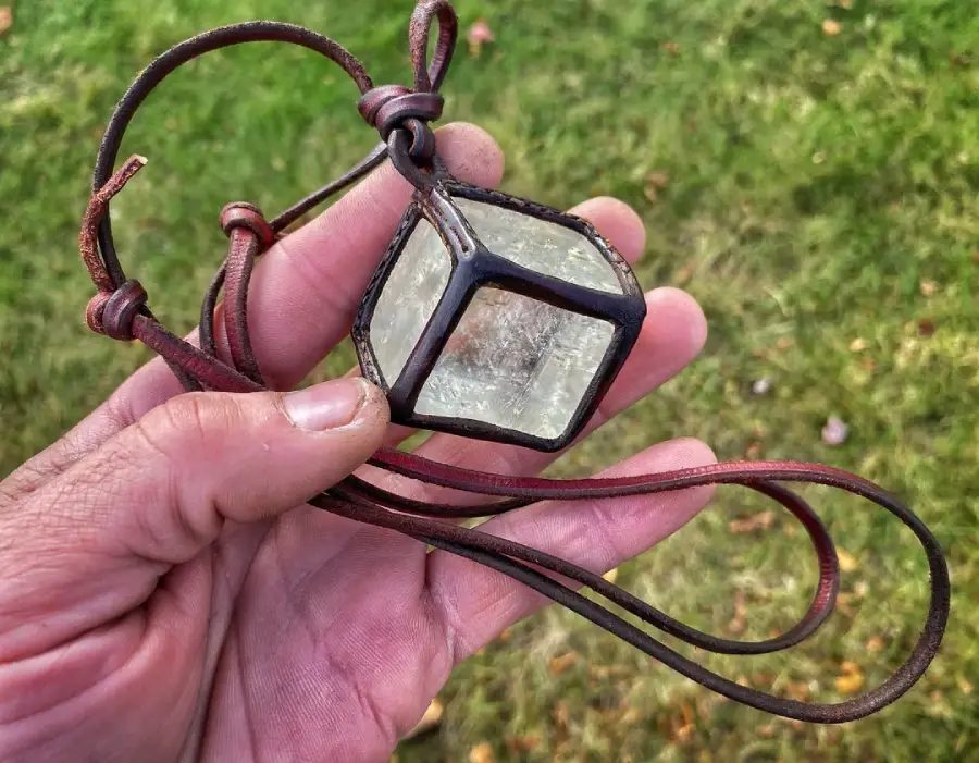 A hand holds a leather cord necklace with a large, clear glass cube pendant encased in a metal frame, shown outdoors over green grass.