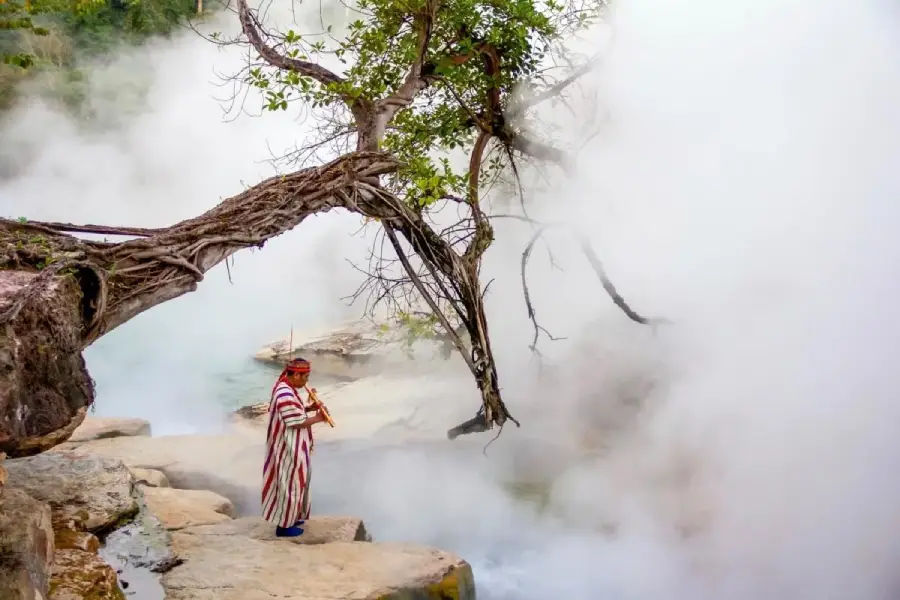 A person wearing a colorful striped robe and a red headband stands on rocks near a steaming river surrounded by mist and an overhanging tree.