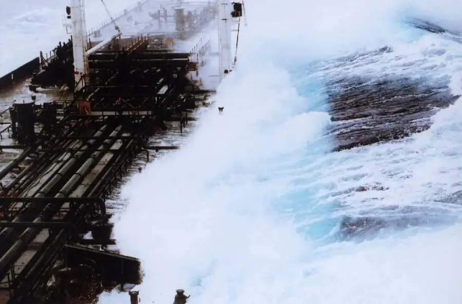 Large ocean waves crash over the deck of a cargo ship, with powerful water surging across the vessel. The sea is rough and turbulent, creating a dramatic contrast between the ship and the churning water.