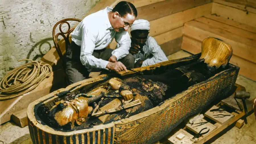 A man examines the golden sarcophagus of an ancient Egyptian mummy, while another person watches closely in the background. The scene appears to take place in an archaeological setting with wooden crates nearby.