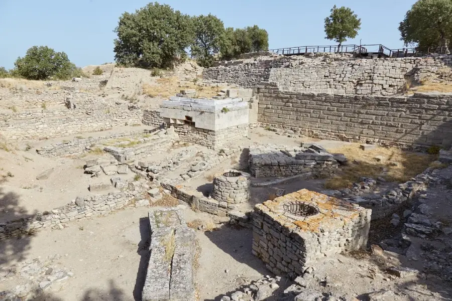 Ancient stone ruins with partially standing walls, a central circular well, and surrounding dry, rocky ground. Sparse grass grows between the stones, and trees dot the background under a clear sky.