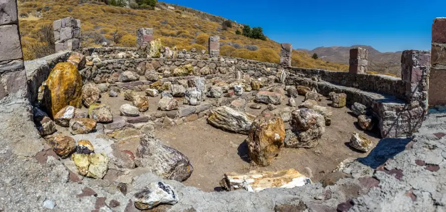 Stone-walled ruins with large rocks scattered within a circular enclosure, set on a dry, hilly landscape under a clear blue sky.