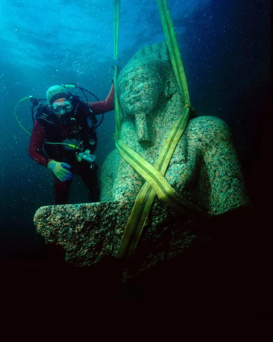 A scuba diver underwater examines a large ancient statue, secured with yellow straps, as it's being lifted from the ocean floor. The statue depicts a seated figure wearing a headdress. Blue water surrounds them.