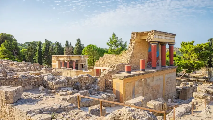Ancient ruins of the Palace of Knossos in Crete, Greece, featuring restored stone walls, red columns, and surrounding green trees under a partly cloudy sky.