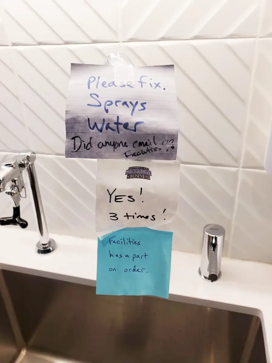 Three notes taped above a sink: "Please fix. Sprays water. Did anyone email facilities?", "Yes! 3 times!", and "Facilities has a part on order." The notes are written in blue and black ink.