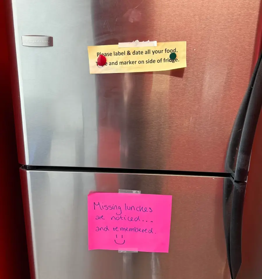 A stainless steel fridge with a yellow note on top reading, "Please label & date all your food, and marker on side of fridge," and a pink note below saying, "Missing lunches are noticed... and remembered! :)