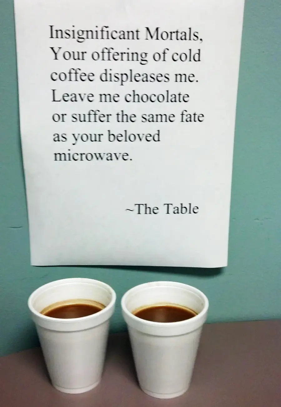 Two white styrofoam cups filled with coffee sit on a table below a humorous sign demanding chocolate and threatening the same fate as a microwave, signed "~The Table.