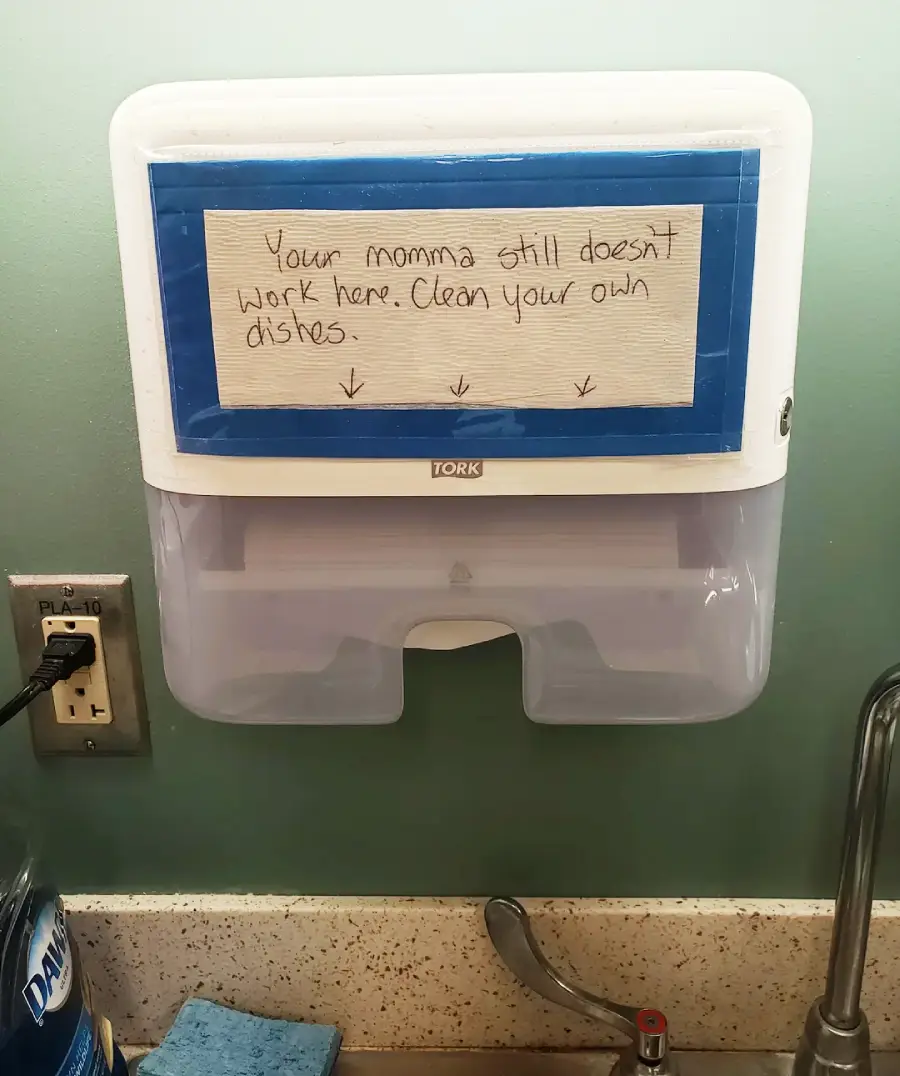A paper towel dispenser on a wall with a handwritten sign that says, "Your momma still doesn’t work here. Clean your own dishes," with arrows pointing down. The dispenser is above a sink and counter with cleaning supplies.