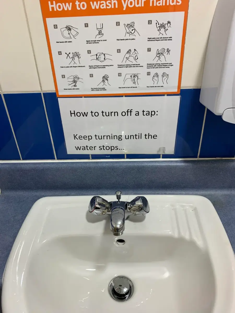 A sink with a silver tap beneath a sign showing handwashing steps and another sign reading, "How to turn off a tap: Keep turning until the water stops..." on a blue and white tiled wall.