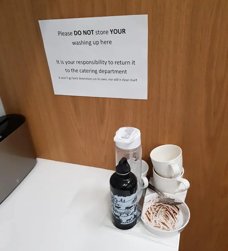 A pile of dirty cups, plates, and bottles stacked on a counter below a sign asking people not to leave their washing up there and to return it to the catering department.
