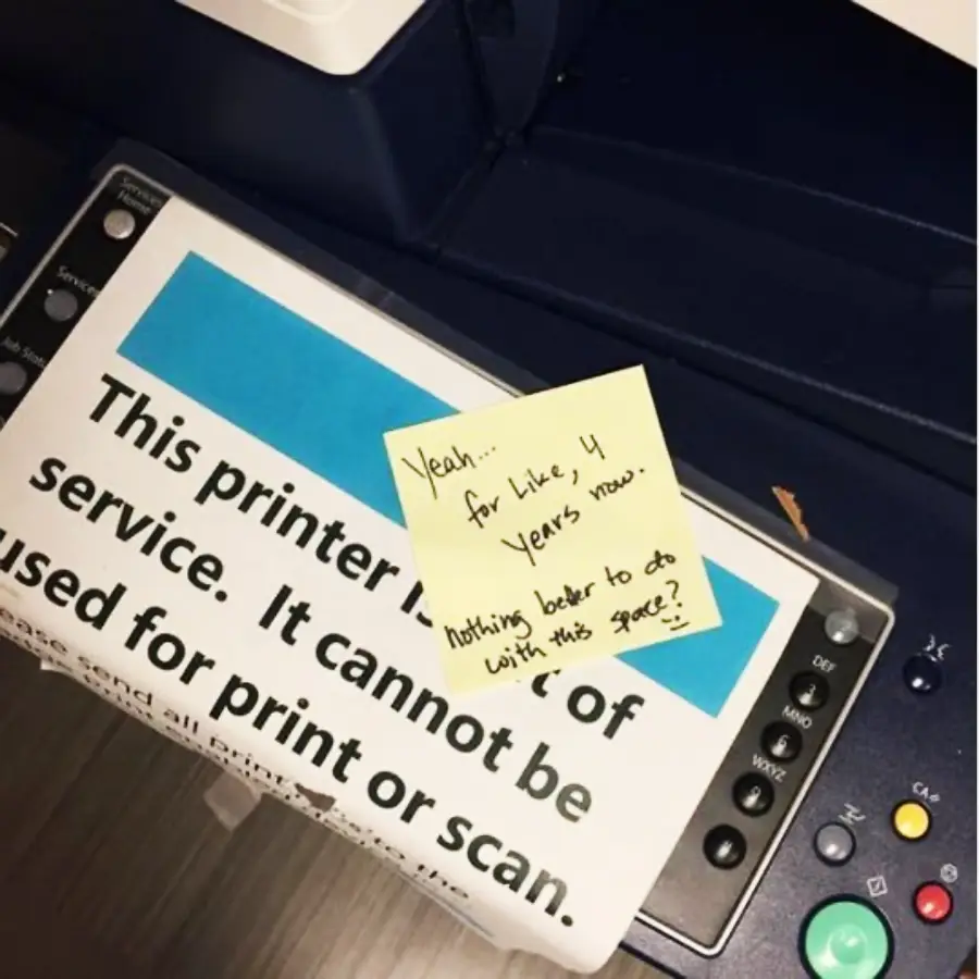 A printer covered with a sign saying, "This printer is out of service. It cannot be used for print or scan." A sticky note on top reads, "Yeah... for like, 4 years now. Nothing better to do with this space?