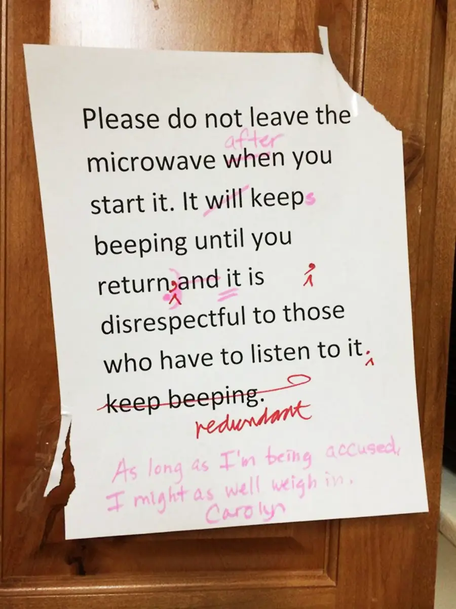 A printed sign with corrected grammar in pink ink reads: "Please do not leave the microwave when you start it. It will keep beeping until you return and it is disrespectful to those who have to listen to it." Handwritten notes correct errors and add, "As long as I'm being accused, I might as well weigh in. Carolyn.