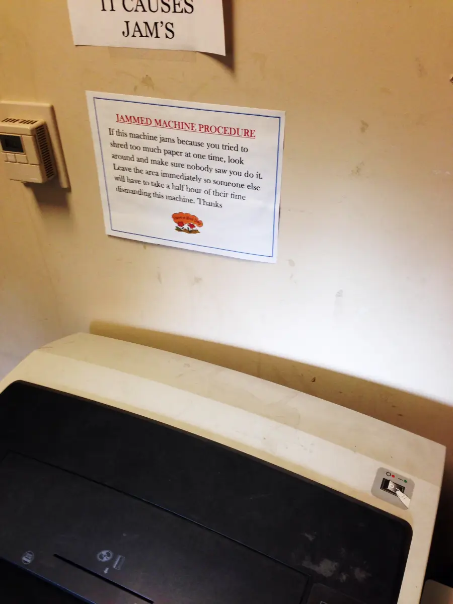 A shredder sits against a beige wall with a printed notice above it titled "Jammed Machine Procedure," giving instructions on what to do if the machine jams due to excess paper.