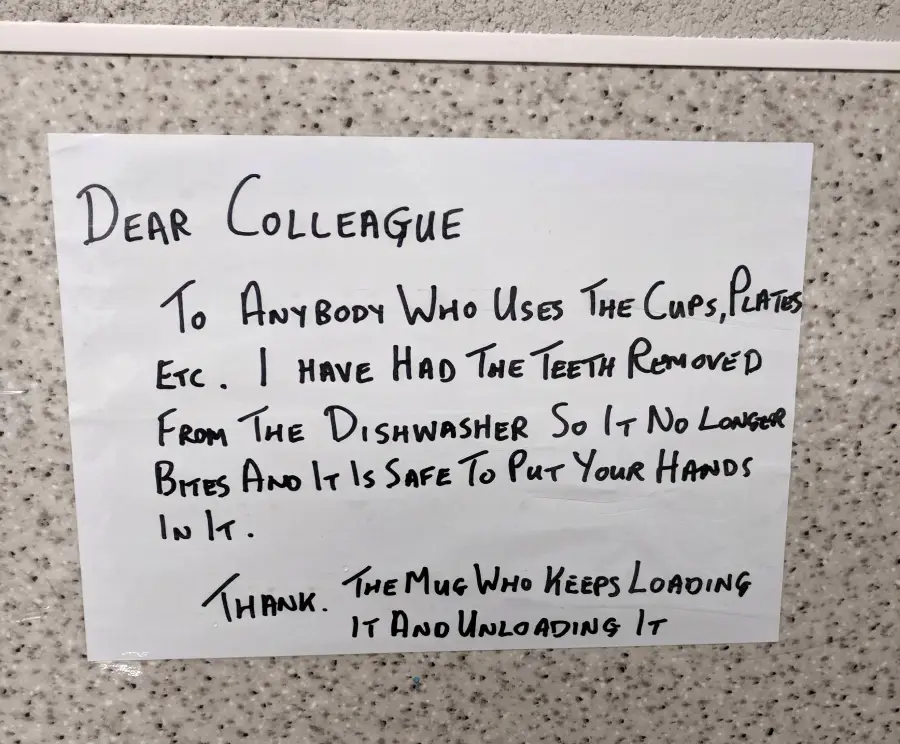 A handwritten sign on a wall reads: "Dear colleague, To anybody who uses the cups, plates etc. I have had the teeth removed from the dishwasher so it no longer bites and it is safe to put your hands in it. Thank the mug who keeps loading it and unloading it.