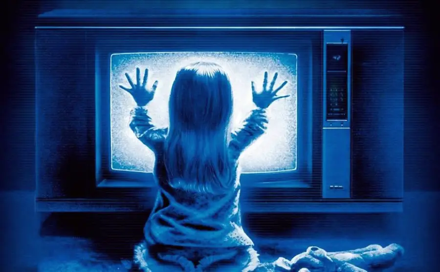A young child in pajamas sits on the floor facing a television screen filled with static, pressing both hands against the glowing screen. The scene is bathed in a blue tint, and a stuffed toy lies nearby.