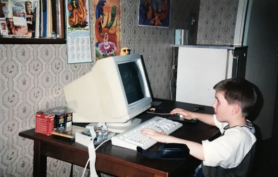 A young boy sits at a desk using an old CRT computer monitor. The room has patterned wallpaper, posters, and shelves with books and floppy disks, evoking a vintage or 1990s atmosphere.