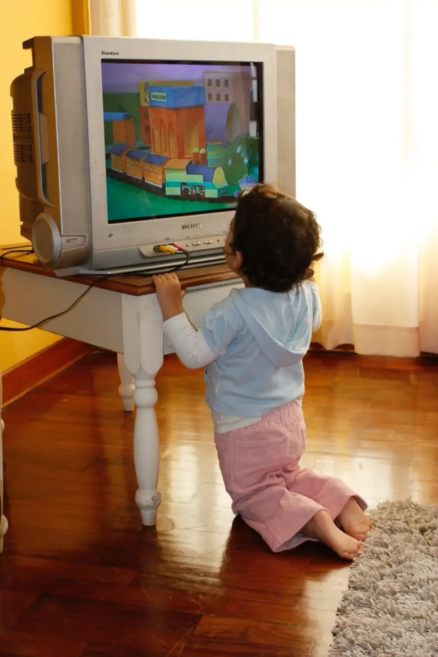 A young child kneels on a wooden floor, closely watching an animated show on a CRT television placed on a small white table in a sunlit room.