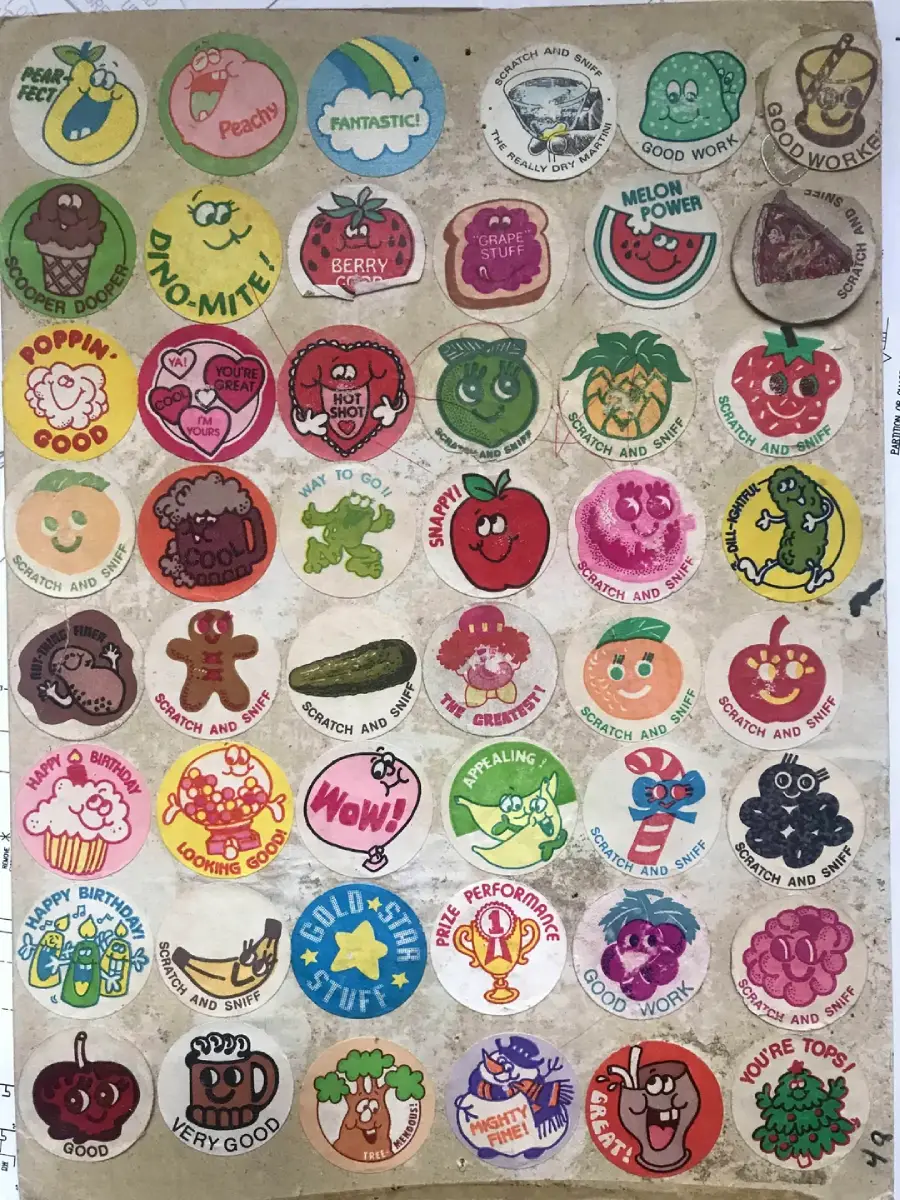 A sheet of colorful, vintage scratch-and-sniff stickers with smiling cartoon fruits, foods, and characters, each with positive messages like "Good Work!" and "Fantastic!" arranged in a grid on a worn backing sheet.