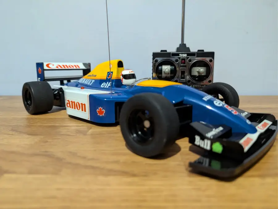 A blue and yellow remote-controlled Formula 1 car sits on a wooden surface, with its controller placed behind it. The car features logos like Canon, elf, and Bull, and has a visible antenna.