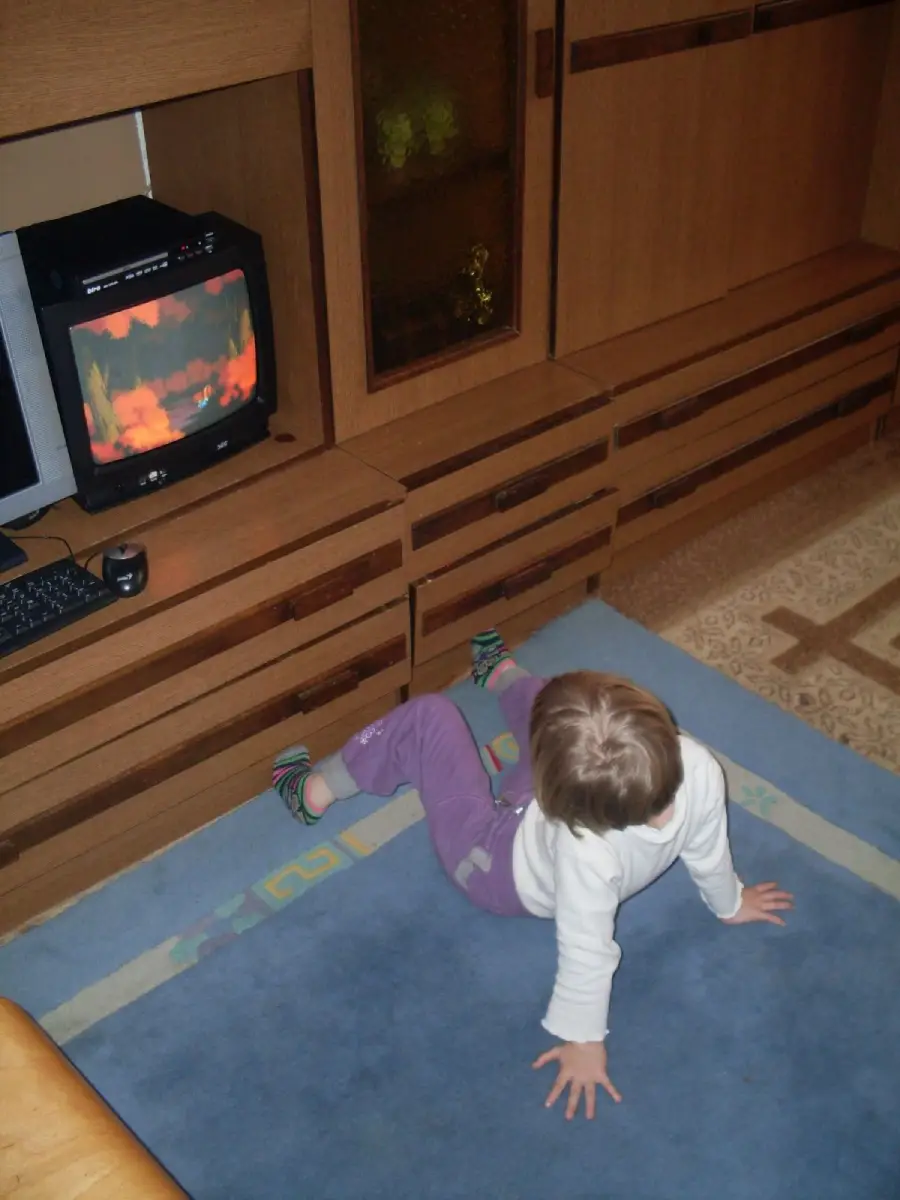 A small child in a white shirt and purple pants sits on a blue rug, leaning back with hands on the floor, watching a TV displaying animated flames in a wood-paneled room.