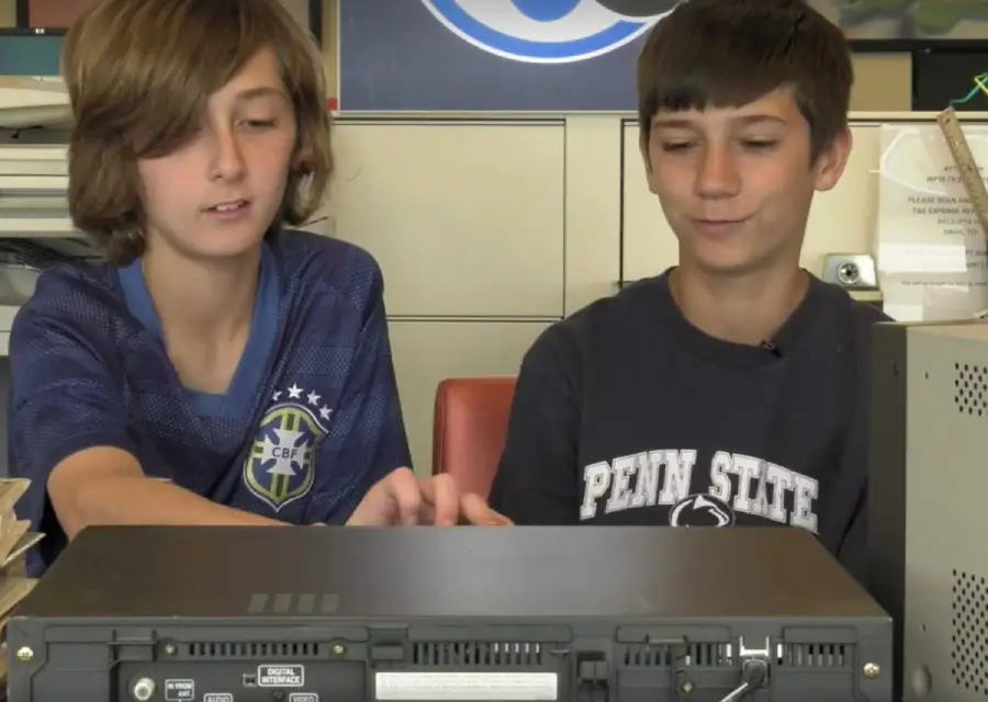 Two boys sit side by side indoors, working together on a piece of electronic equipment. One wears a blue soccer jersey, the other a dark Penn State shirt. Both are focused on the device in front of them.