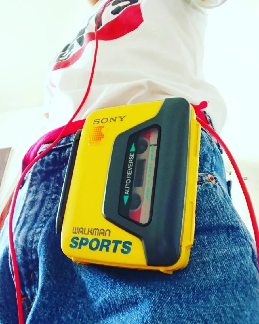 Close-up of a person wearing blue jeans and a white shirt, with a yellow Sony Walkman Sports cassette player clipped to their waistband and red earphone cables hanging down.
